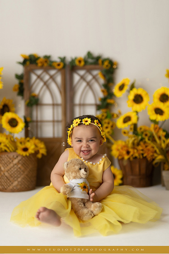 First birthday sunflower cake smash photography session featuring a baby girl in a yellow dress and sunflower headband, surrounded by bright sunflowers, rustic wood details, a teddy bear, and a yellow and white birthday cake at Studio 120 Photography in Ramona, California.