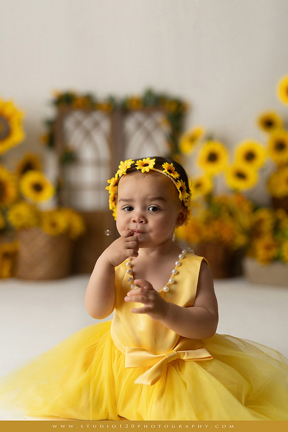 First birthday sunflower cake smash photography session featuring a baby girl in a yellow dress and sunflower headband, surrounded by bright sunflowers, rustic wood details, a teddy bear, and a yellow and white birthday cake at Studio 120 Photography in Ramona, California.
