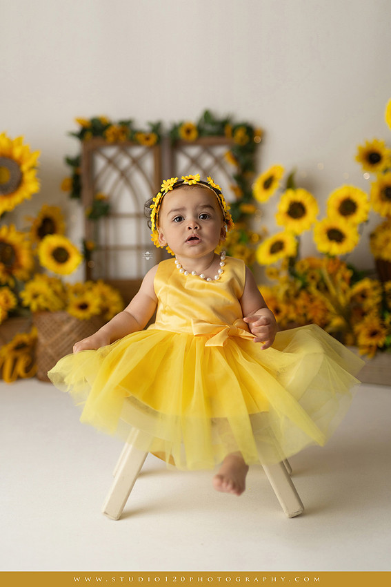 First birthday sunflower cake smash photography session featuring a baby girl in a yellow dress and sunflower headband, surrounded by bright sunflowers, rustic wood details, a teddy bear, and a yellow and white birthday cake at Studio 120 Photography in Ramona, California.