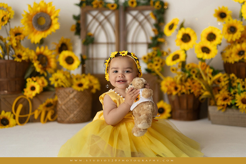 First birthday sunflower cake smash photography session featuring a baby girl in a yellow dress and sunflower headband, surrounded by bright sunflowers, rustic wood details, a teddy bear, and a yellow and white birthday cake at Studio 120 Photography in Ramona, California.