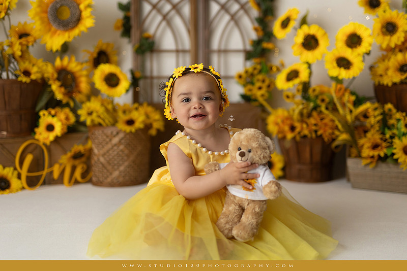 First birthday sunflower cake smash photography session featuring a baby girl in a yellow dress and sunflower headband, surrounded by bright sunflowers, rustic wood details, a teddy bear, and a yellow and white birthday cake at Studio 120 Photography in Ramona, California.