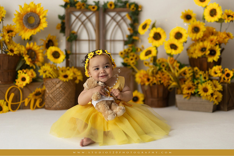 First birthday sunflower cake smash photography session featuring a baby girl in a yellow dress and sunflower headband, surrounded by bright sunflowers, rustic wood details, a teddy bear, and a yellow and white birthday cake at Studio 120 Photography in Ramona, California.