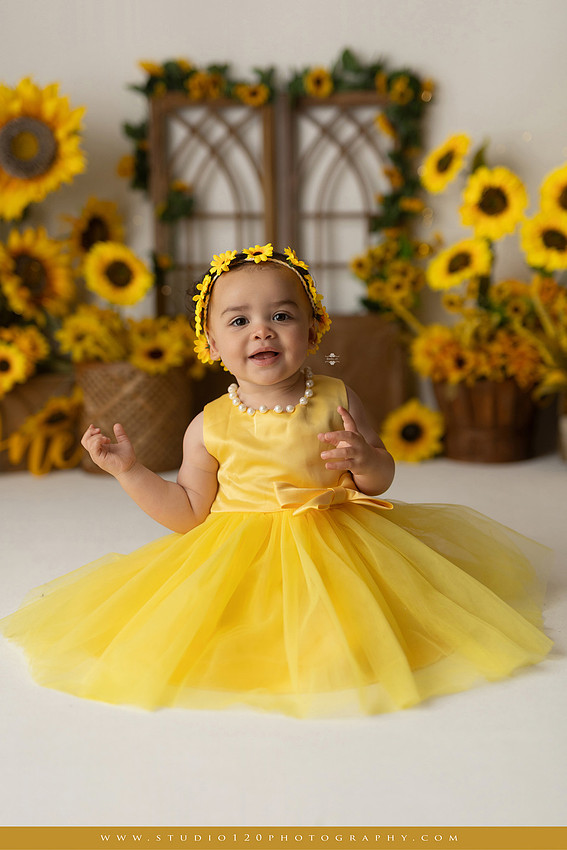 First birthday sunflower cake smash photography session featuring a baby girl in a yellow dress and sunflower headband, surrounded by bright sunflowers, rustic wood details, a teddy bear, and a yellow and white birthday cake at Studio 120 Photography in Ramona, California.