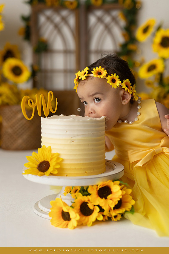 First birthday sunflower cake smash photography session featuring a baby girl in a yellow dress and sunflower headband, surrounded by bright sunflowers, rustic wood details, a teddy bear, and a yellow and white birthday cake at Studio 120 Photography in Ramona, California.