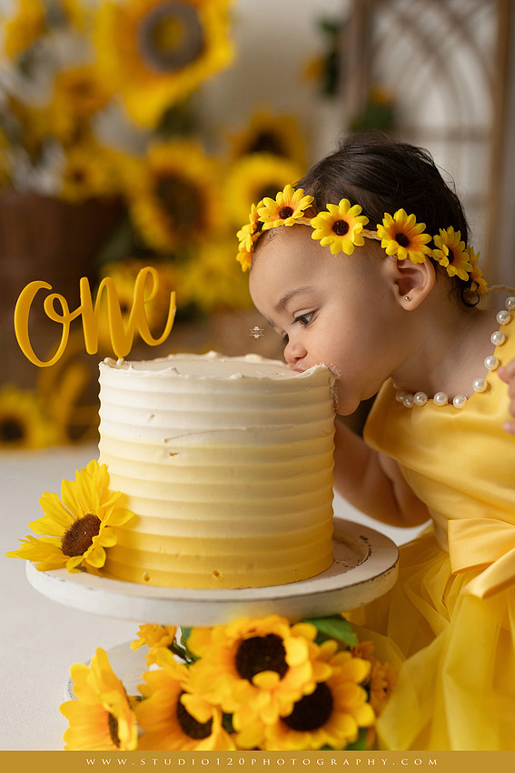 First birthday sunflower cake smash photography session featuring a baby girl in a yellow dress and sunflower headband, surrounded by bright sunflowers, rustic wood details, a teddy bear, and a yellow and white birthday cake at Studio 120 Photography in Ramona, California.