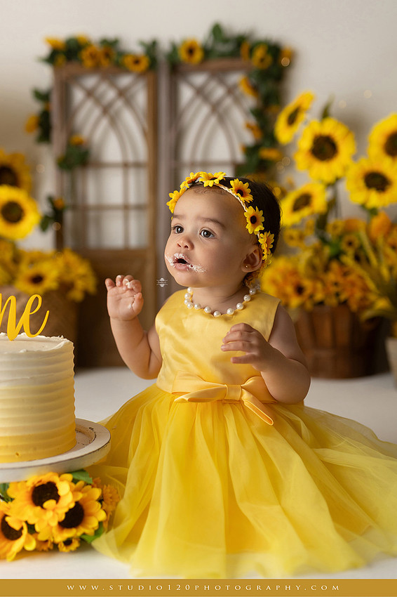 First birthday sunflower cake smash photography session featuring a baby girl in a yellow dress and sunflower headband, surrounded by bright sunflowers, rustic wood details, a teddy bear, and a yellow and white birthday cake at Studio 120 Photography in Ramona, California.