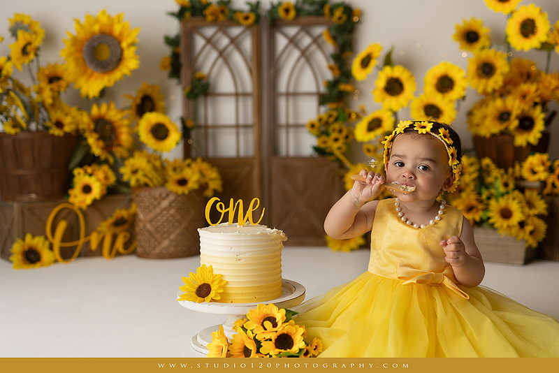 First birthday sunflower cake smash photography session featuring a baby girl in a yellow dress and sunflower headband, surrounded by bright sunflowers, rustic wood details, a teddy bear, and a yellow and white birthday cake at Studio 120 Photography in Ramona, California.