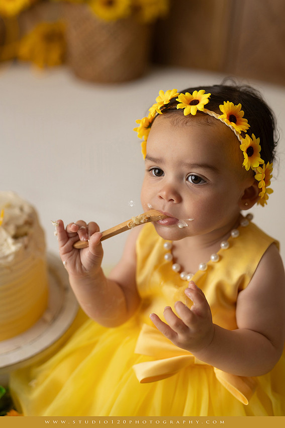 First birthday sunflower cake smash photography session featuring a baby girl in a yellow dress and sunflower headband, surrounded by bright sunflowers, rustic wood details, a teddy bear, and a yellow and white birthday cake at Studio 120 Photography in Ramona, California.