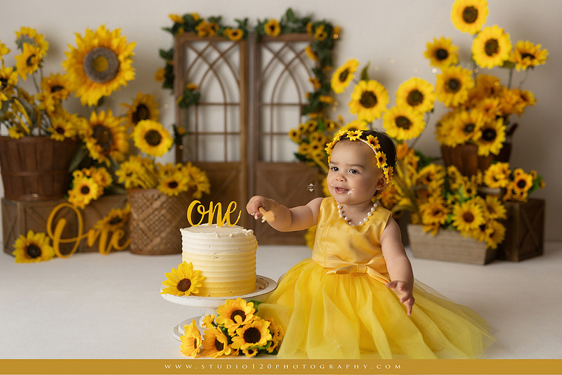 First birthday sunflower cake smash photography session featuring a baby girl in a yellow dress and sunflower headband, surrounded by bright sunflowers, rustic wood details, a teddy bear, and a yellow and white birthday cake at Studio 120 Photography in Ramona, California.