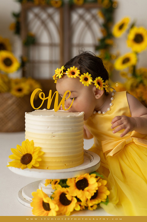 First birthday sunflower cake smash photography session featuring a baby girl in a yellow dress and sunflower headband, surrounded by bright sunflowers, rustic wood details, a teddy bear, and a yellow and white birthday cake at Studio 120 Photography in Ramona, California.