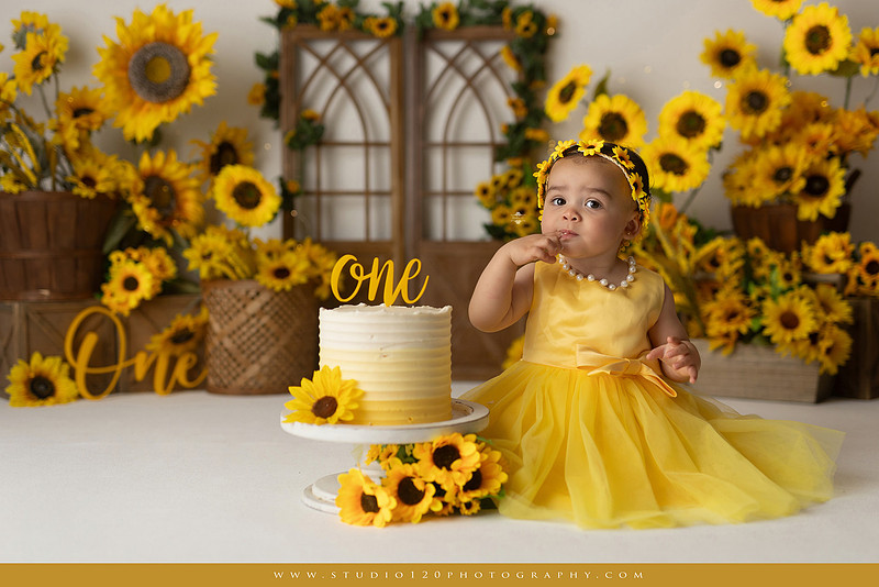 First birthday sunflower cake smash photography session featuring a baby girl in a yellow dress and sunflower headband, surrounded by bright sunflowers, rustic wood details, a teddy bear, and a yellow and white birthday cake at Studio 120 Photography in Ramona, California.