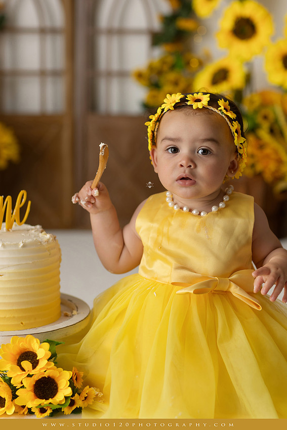 First birthday sunflower cake smash photography session featuring a baby girl in a yellow dress and sunflower headband, surrounded by bright sunflowers, rustic wood details, a teddy bear, and a yellow and white birthday cake at Studio 120 Photography in Ramona, California.