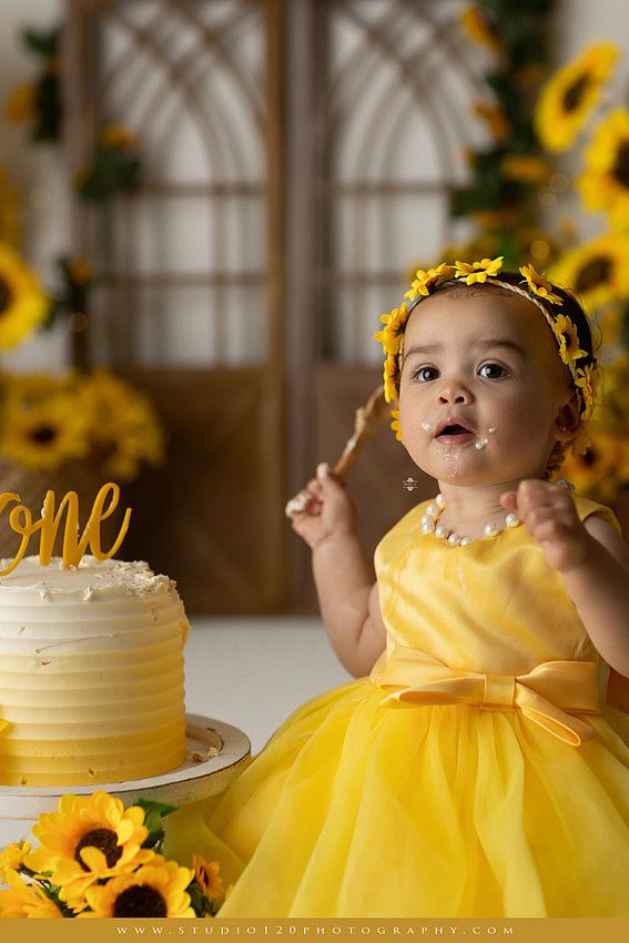 First birthday sunflower cake smash photography session featuring a baby girl in a yellow dress and sunflower headband, surrounded by bright sunflowers, rustic wood details, a teddy bear, and a yellow and white birthday cake at Studio 120 Photography in Ramona, California.