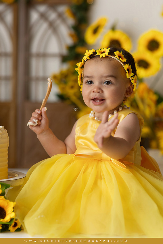 First birthday sunflower cake smash photography session featuring a baby girl in a yellow dress and sunflower headband, surrounded by bright sunflowers, rustic wood details, a teddy bear, and a yellow and white birthday cake at Studio 120 Photography in Ramona, California.