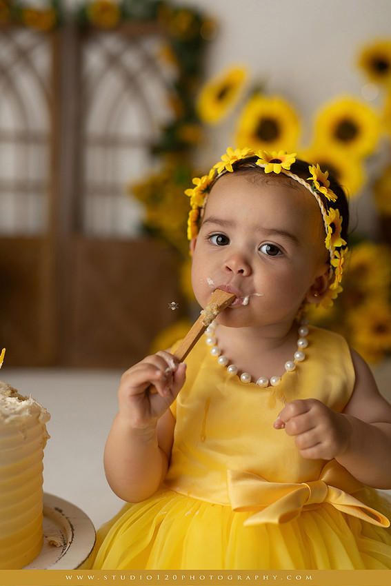 First birthday sunflower cake smash photography session featuring a baby girl in a yellow dress and sunflower headband, surrounded by bright sunflowers, rustic wood details, a teddy bear, and a yellow and white birthday cake at Studio 120 Photography in Ramona, California.