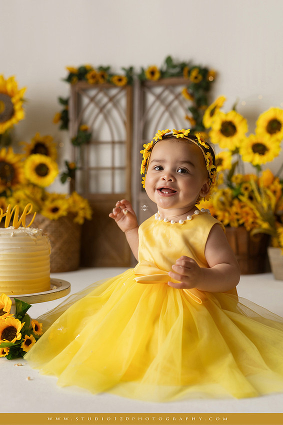 First birthday sunflower cake smash photography session featuring a baby girl in a yellow dress and sunflower headband, surrounded by bright sunflowers, rustic wood details, a teddy bear, and a yellow and white birthday cake at Studio 120 Photography in Ramona, California.