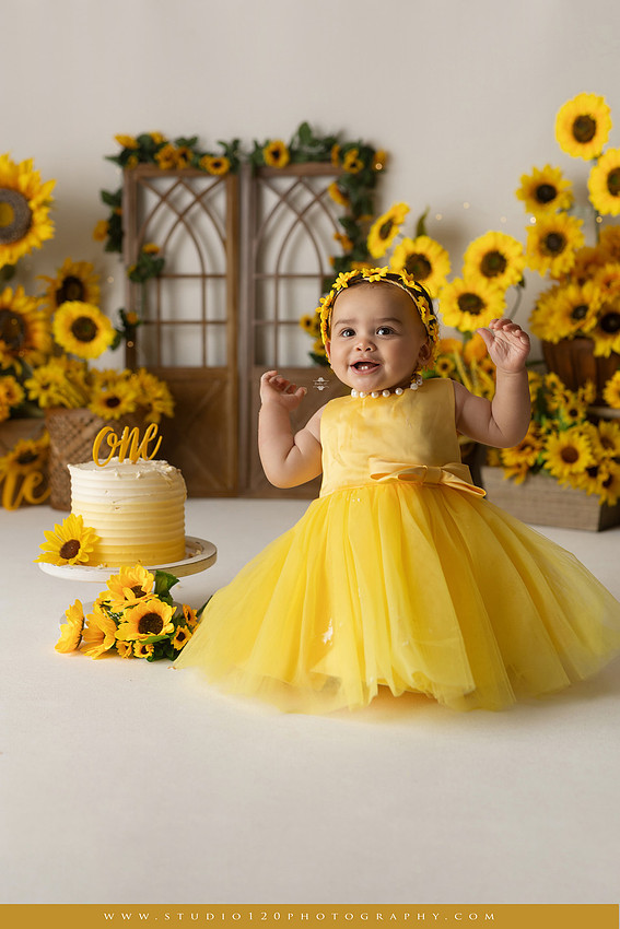First birthday sunflower cake smash photography session featuring a baby girl in a yellow dress and sunflower headband, surrounded by bright sunflowers, rustic wood details, a teddy bear, and a yellow and white birthday cake at Studio 120 Photography in Ramona, California.