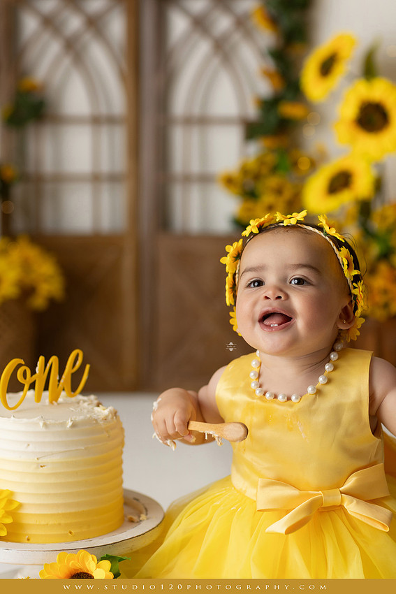 First birthday sunflower cake smash photography session featuring a baby girl in a yellow dress and sunflower headband, surrounded by bright sunflowers, rustic wood details, a teddy bear, and a yellow and white birthday cake at Studio 120 Photography in Ramona, California.