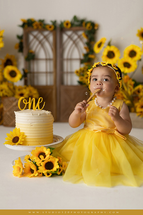First birthday sunflower cake smash photography session featuring a baby girl in a yellow dress and sunflower headband, surrounded by bright sunflowers, rustic wood details, a teddy bear, and a yellow and white birthday cake at Studio 120 Photography in Ramona, California.