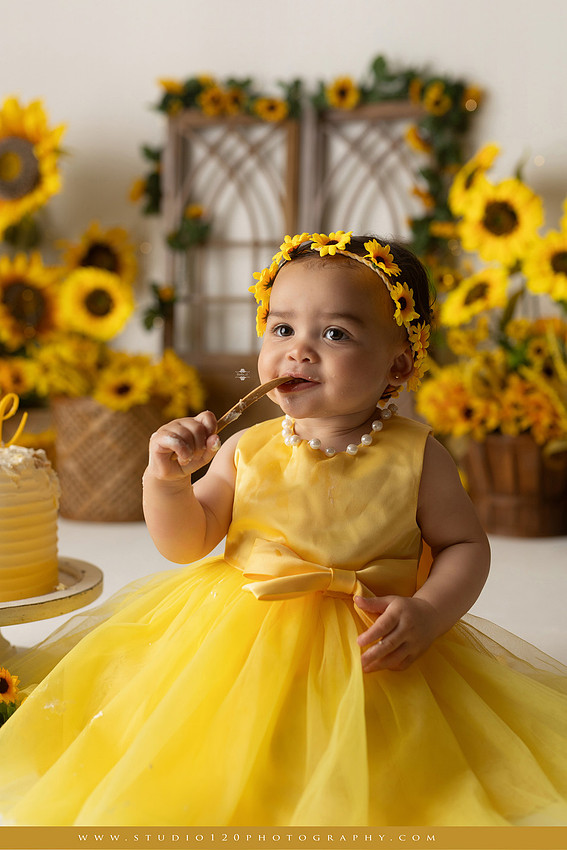 First birthday sunflower cake smash photography session featuring a baby girl in a yellow dress and sunflower headband, surrounded by bright sunflowers, rustic wood details, a teddy bear, and a yellow and white birthday cake at Studio 120 Photography in Ramona, California.