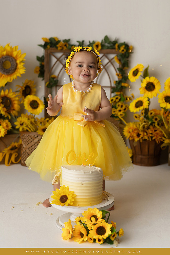 First birthday sunflower cake smash photography session featuring a baby girl in a yellow dress and sunflower headband, surrounded by bright sunflowers, rustic wood details, a teddy bear, and a yellow and white birthday cake at Studio 120 Photography in Ramona, California.