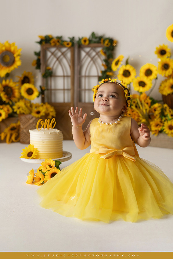 First birthday sunflower cake smash photography session featuring a baby girl in a yellow dress and sunflower headband, surrounded by bright sunflowers, rustic wood details, a teddy bear, and a yellow and white birthday cake at Studio 120 Photography in Ramona, California.
