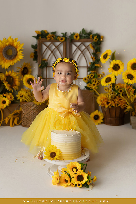 First birthday sunflower cake smash photography session featuring a baby girl in a yellow dress and sunflower headband, surrounded by bright sunflowers, rustic wood details, a teddy bear, and a yellow and white birthday cake at Studio 120 Photography in Ramona, California.