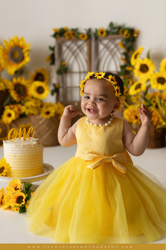 First birthday sunflower cake smash photography session featuring a baby girl in a yellow dress and sunflower headband, surrounded by bright sunflowers, rustic wood details, a teddy bear, and a yellow and white birthday cake at Studio 120 Photography in Ramona, California.