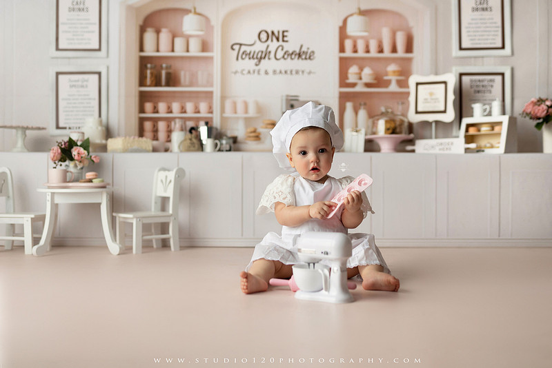 One Tough Cookie first birthday cake smash photography session featuring a baby girl in a soft pink bakery-themed set, chef outfit, floral dress, cookies, birthday cake and cake smash portraits at Studio 120 Photography in Ramona, California.