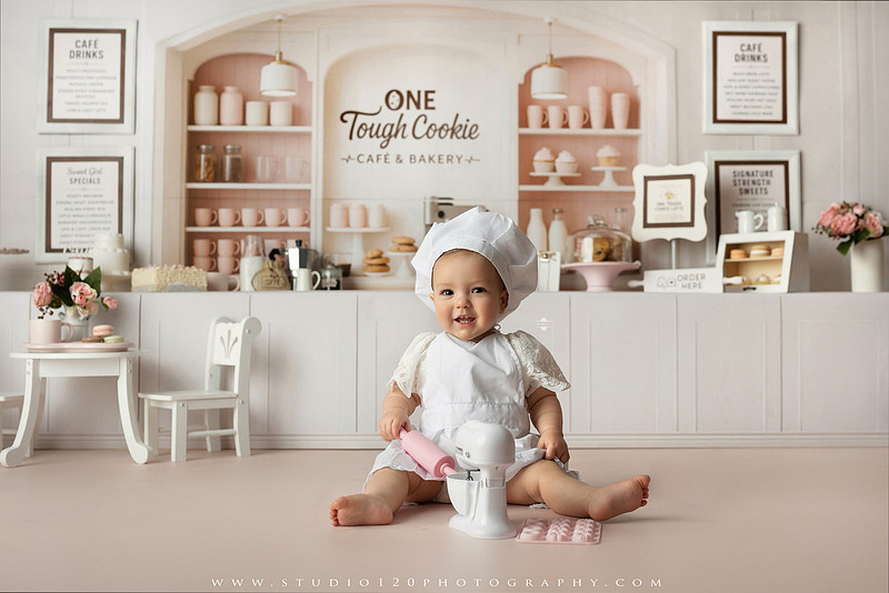 One Tough Cookie first birthday cake smash photography session featuring a baby girl in a soft pink bakery-themed set, chef outfit, floral dress, cookies, birthday cake and cake smash portraits at Studio 120 Photography in Ramona, California.