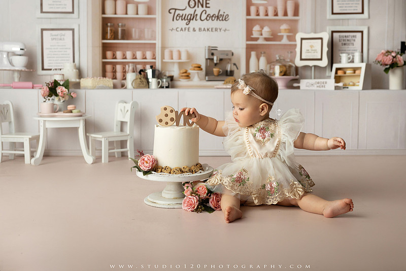 One Tough Cookie first birthday cake smash photography session featuring a baby girl in a soft pink bakery-themed set, chef outfit, floral dress, cookies, birthday cake and cake smash portraits at Studio 120 Photography in Ramona, California.