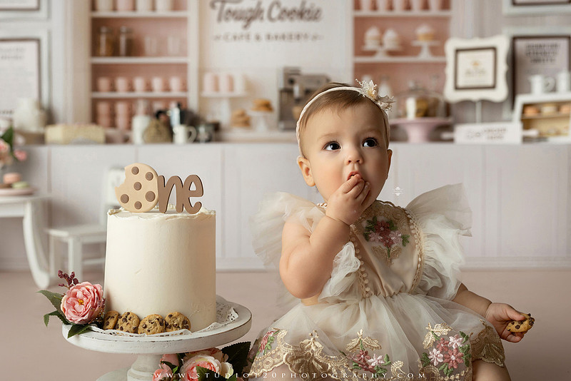 One Tough Cookie first birthday cake smash photography session featuring a baby girl in a soft pink bakery-themed set, chef outfit, floral dress, cookies, birthday cake and cake smash portraits at Studio 120 Photography in Ramona, California.