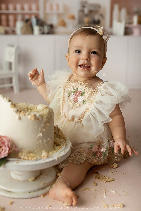 One Tough Cookie first birthday cake smash photography session featuring a baby girl in a soft pink bakery-themed set, chef outfit, floral dress, cookies, birthday cake and cake smash portraits at Studio 120 Photography in Ramona, California.