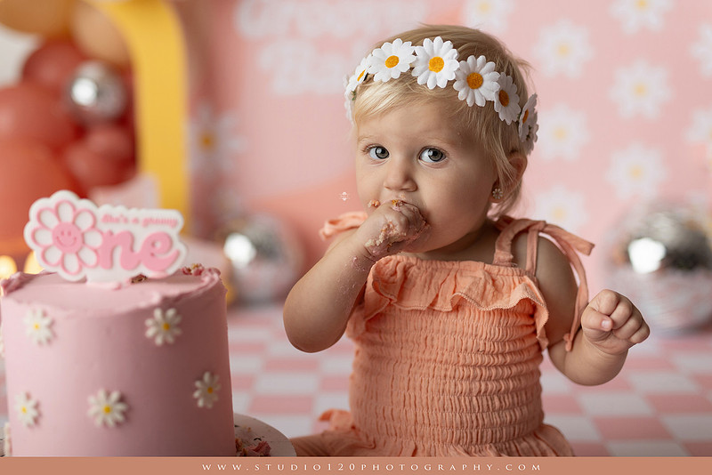 One Groovy Babe first birthday cake smash photography session at Studio 120 Photography in Ramona, California, featuring a baby girl in a pink and yellow retro daisy and disco themed set with balloons, a pink birthday cake, flower crown and vintage groovy van, followed by a rustic apple milk bath with red apples, florals and wooden accents.