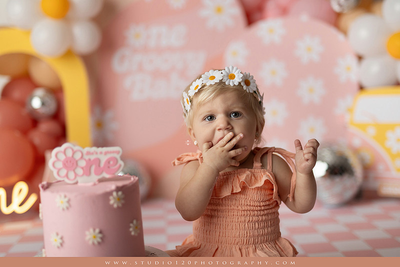 One Groovy Babe first birthday cake smash photography session at Studio 120 Photography in Ramona, California, featuring a baby girl in a pink and yellow retro daisy and disco themed set with balloons, a pink birthday cake, flower crown and vintage groovy van, followed by a rustic apple milk bath with red apples, florals and wooden accents.