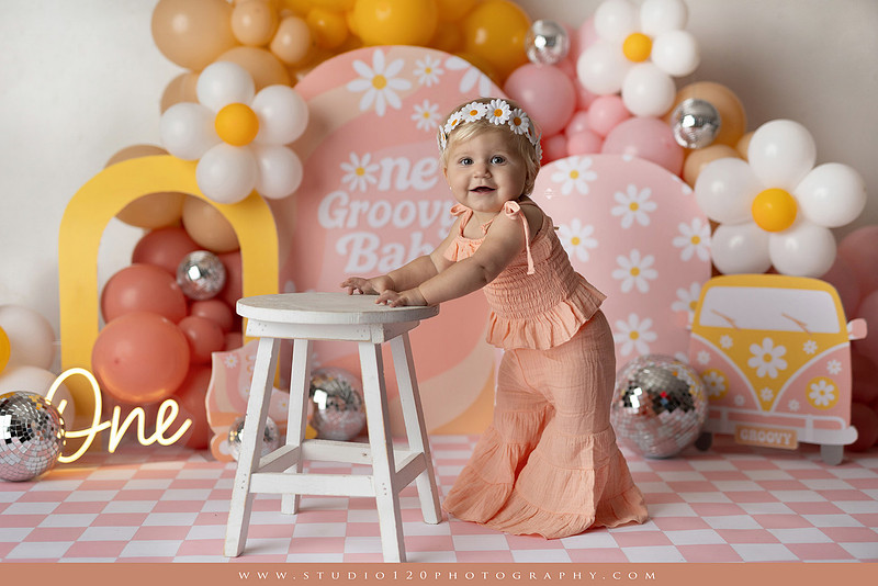 One Groovy Babe first birthday cake smash photography session at Studio 120 Photography in Ramona, California, featuring a baby girl in a pink and yellow retro daisy and disco themed set with balloons, a pink birthday cake, flower crown and vintage groovy van, followed by a rustic apple milk bath with red apples, florals and wooden accents.