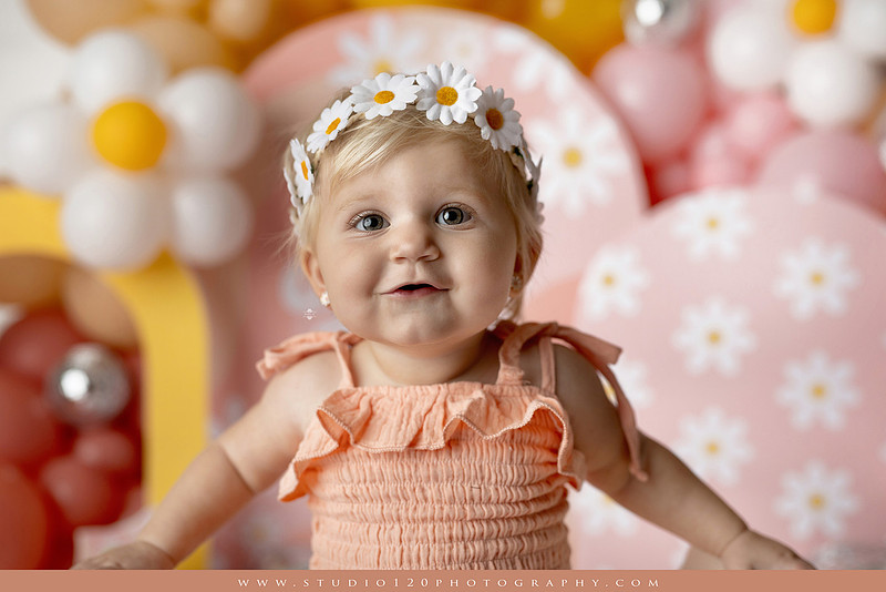 One Groovy Babe first birthday cake smash photography session at Studio 120 Photography in Ramona, California, featuring a baby girl in a pink and yellow retro daisy and disco themed set with balloons, a pink birthday cake, flower crown and vintage groovy van, followed by a rustic apple milk bath with red apples, florals and wooden accents.