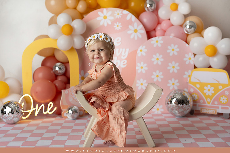 One Groovy Babe first birthday cake smash photography session at Studio 120 Photography in Ramona, California, featuring a baby girl in a pink and yellow retro daisy and disco themed set with balloons, a pink birthday cake, flower crown and vintage groovy van, followed by a rustic apple milk bath with red apples, florals and wooden accents.