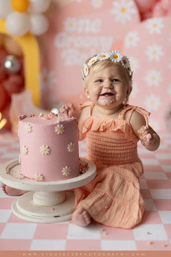 One Groovy Babe first birthday cake smash photography session at Studio 120 Photography in Ramona, California, featuring a baby girl in a pink and yellow retro daisy and disco themed set with balloons, a pink birthday cake, flower crown and vintage groovy van, followed by a rustic apple milk bath with red apples, florals and wooden accents.