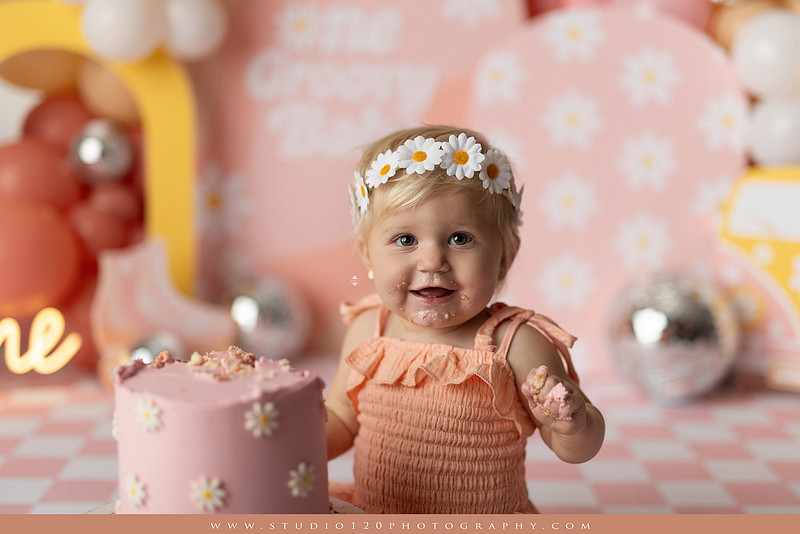 One Groovy Babe first birthday cake smash photography session at Studio 120 Photography in Ramona, California, featuring a baby girl in a pink and yellow retro daisy and disco themed set with balloons, a pink birthday cake, flower crown and vintage groovy van, followed by a rustic apple milk bath with red apples, florals and wooden accents.