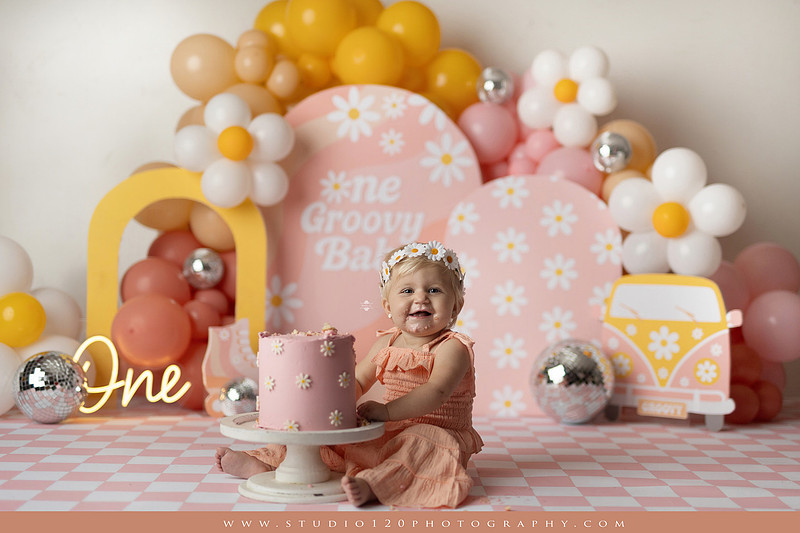 One Groovy Babe first birthday cake smash photography session at Studio 120 Photography in Ramona, California, featuring a baby girl in a pink and yellow retro daisy and disco themed set with balloons, a pink birthday cake, flower crown and vintage groovy van, followed by a rustic apple milk bath with red apples, florals and wooden accents.