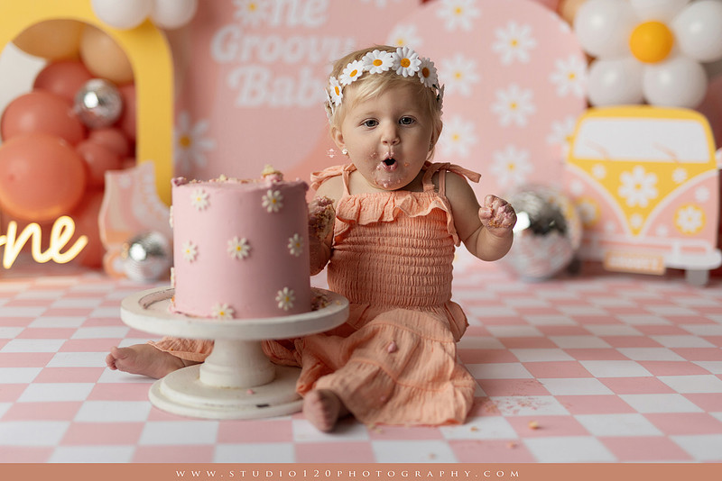 One Groovy Babe first birthday cake smash photography session at Studio 120 Photography in Ramona, California, featuring a baby girl in a pink and yellow retro daisy and disco themed set with balloons, a pink birthday cake, flower crown and vintage groovy van, followed by a rustic apple milk bath with red apples, florals and wooden accents.