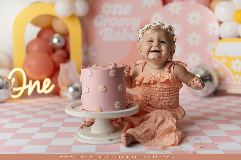 One Groovy Babe first birthday cake smash photography session at Studio 120 Photography in Ramona, California, featuring a baby girl in a pink and yellow retro daisy and disco themed set with balloons, a pink birthday cake, flower crown and vintage groovy van, followed by a rustic apple milk bath with red apples, florals and wooden accents.