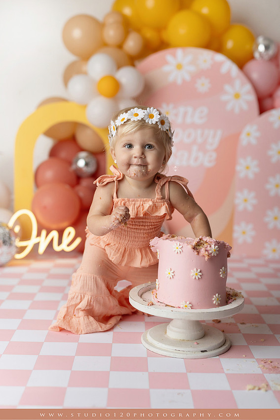 One Groovy Babe first birthday cake smash photography session at Studio 120 Photography in Ramona, California, featuring a baby girl in a pink and yellow retro daisy and disco themed set with balloons, a pink birthday cake, flower crown and vintage groovy van, followed by a rustic apple milk bath with red apples, florals and wooden accents.