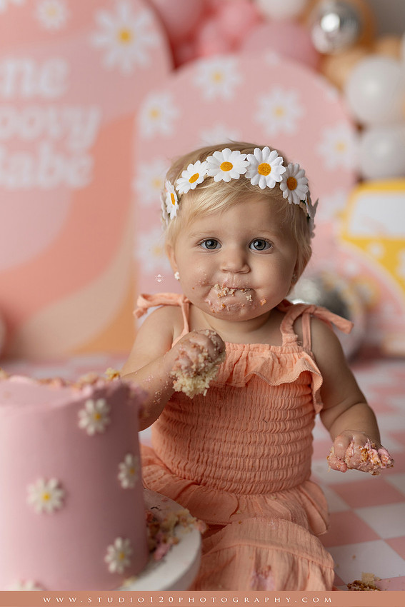 One Groovy Babe first birthday cake smash photography session at Studio 120 Photography in Ramona, California, featuring a baby girl in a pink and yellow retro daisy and disco themed set with balloons, a pink birthday cake, flower crown and vintage groovy van, followed by a rustic apple milk bath with red apples, florals and wooden accents.