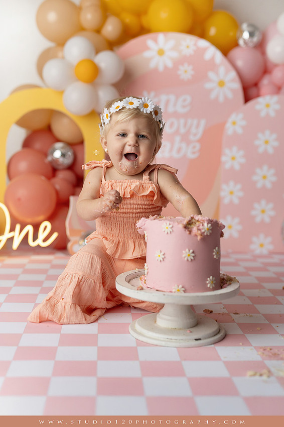 One Groovy Babe first birthday cake smash photography session at Studio 120 Photography in Ramona, California, featuring a baby girl in a pink and yellow retro daisy and disco themed set with balloons, a pink birthday cake, flower crown and vintage groovy van, followed by a rustic apple milk bath with red apples, florals and wooden accents.