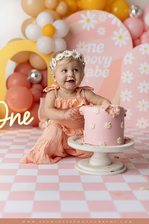 One Groovy Babe first birthday cake smash photography session at Studio 120 Photography in Ramona, California, featuring a baby girl in a pink and yellow retro daisy and disco themed set with balloons, a pink birthday cake, flower crown and vintage groovy van, followed by a rustic apple milk bath with red apples, florals and wooden accents.