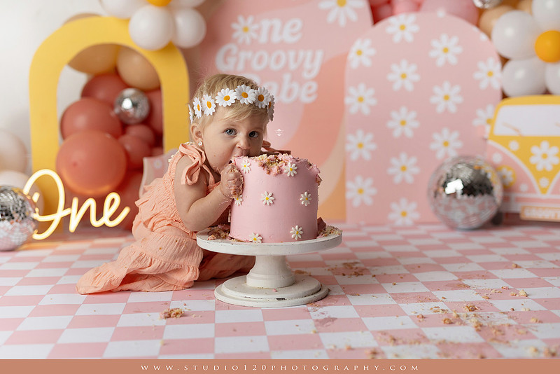 One Groovy Babe first birthday cake smash photography session at Studio 120 Photography in Ramona, California, featuring a baby girl in a pink and yellow retro daisy and disco themed set with balloons, a pink birthday cake, flower crown and vintage groovy van, followed by a rustic apple milk bath with red apples, florals and wooden accents.