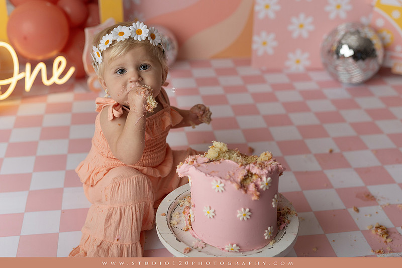 One Groovy Babe first birthday cake smash photography session at Studio 120 Photography in Ramona, California, featuring a baby girl in a pink and yellow retro daisy and disco themed set with balloons, a pink birthday cake, flower crown and vintage groovy van, followed by a rustic apple milk bath with red apples, florals and wooden accents.