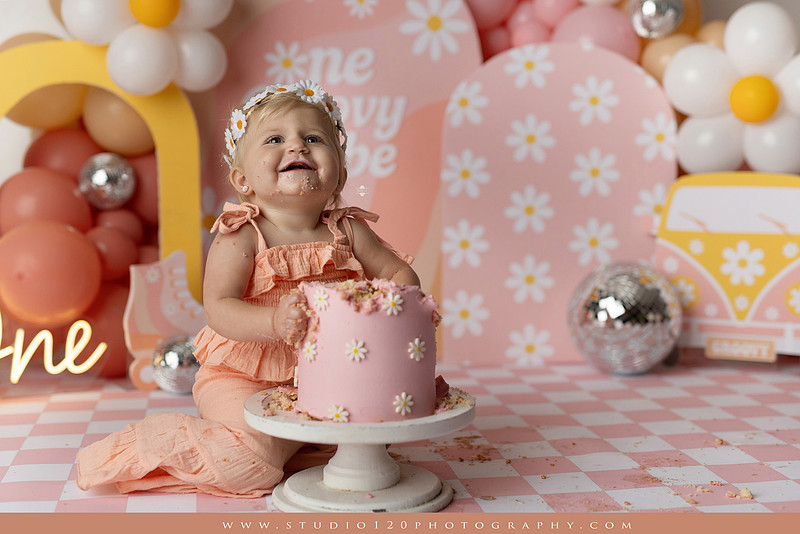 One Groovy Babe first birthday cake smash photography session at Studio 120 Photography in Ramona, California, featuring a baby girl in a pink and yellow retro daisy and disco themed set with balloons, a pink birthday cake, flower crown and vintage groovy van, followed by a rustic apple milk bath with red apples, florals and wooden accents.